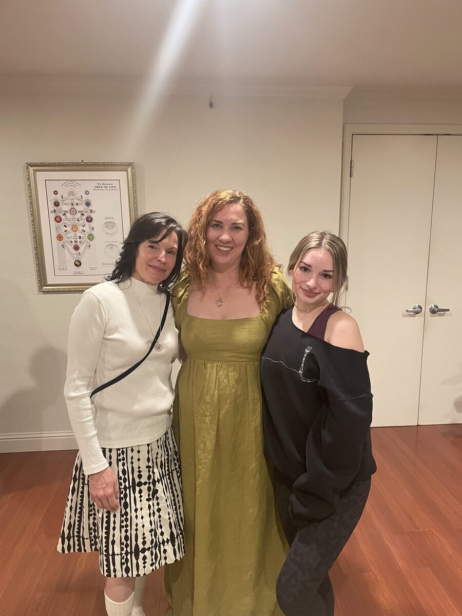 Izzy with two women at a gathering, a framed Tree of Life on the wall behind them